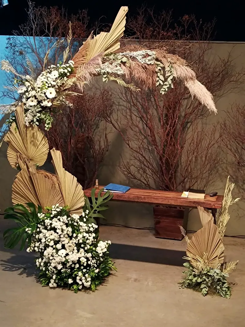 Arco de folhas de palmeira secas, plumas de capim e flores brancas, com galhos secos e um banco de madeira
