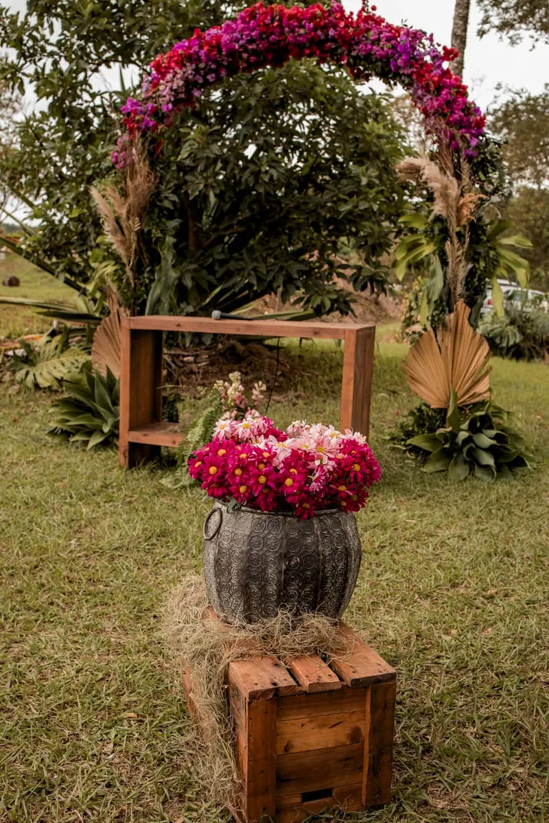 Arco de flores rosa e roxas no gramado, com um banco de madeira atrás e um vaso de flores sobre caixotes