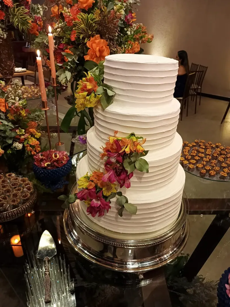 Bolo branco de andares, com flores laranjas, num suporte prateado entre velas