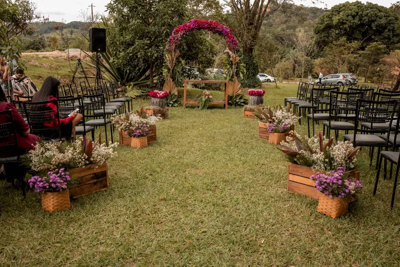 Cerimônia ao ar livre no gramado, com cadeiras pretas, caixotes de madeira com flores e um arco de flores coloridas