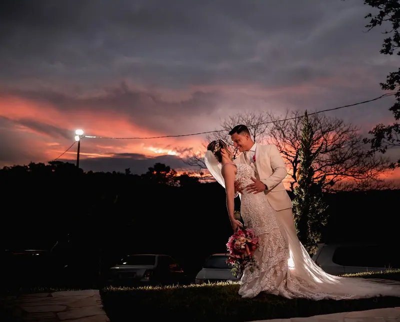 Noivos abraçados no gramado ao pôr do sol, a noiva de vestido de renda e cauda longa com um buquê de flores rosa, o noivo de terno claro, céu escuro com nuvens alaranjadas e árvores em silhueta