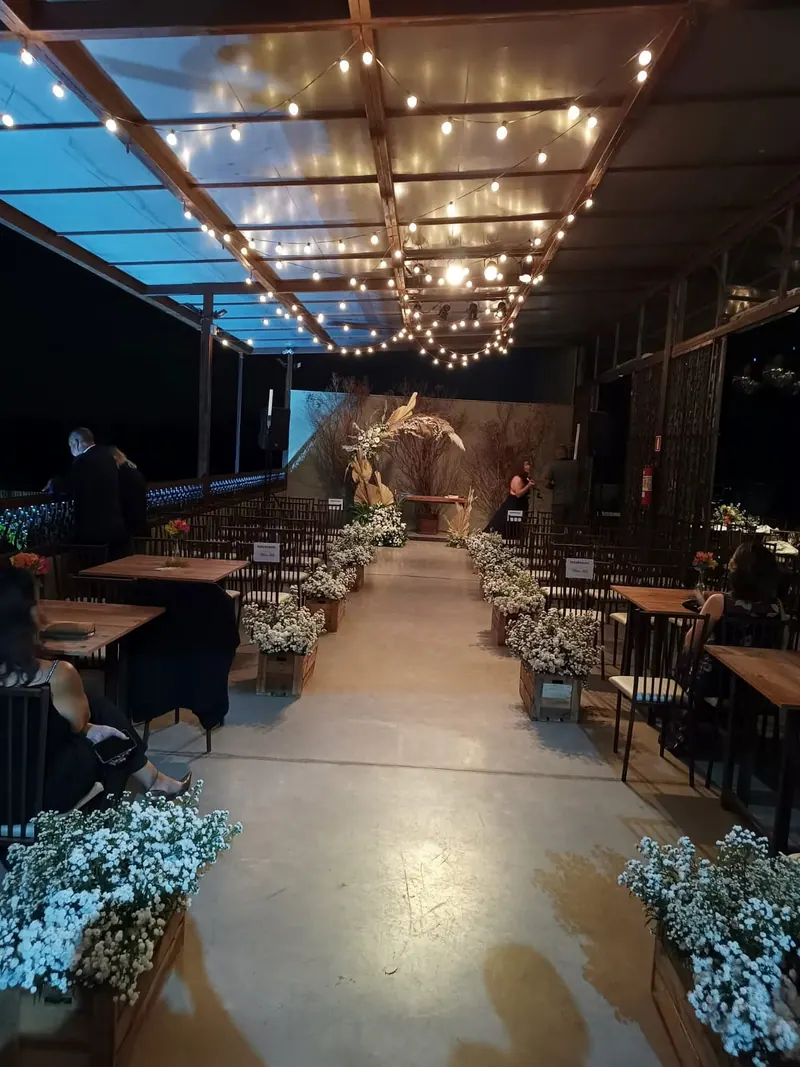 Salão coberto à noite, com fios de lâmpadas no teto, um corredor de cerimônia entre caixotes de madeira com flores brancas e, ao fundo, um arco de folhas secas